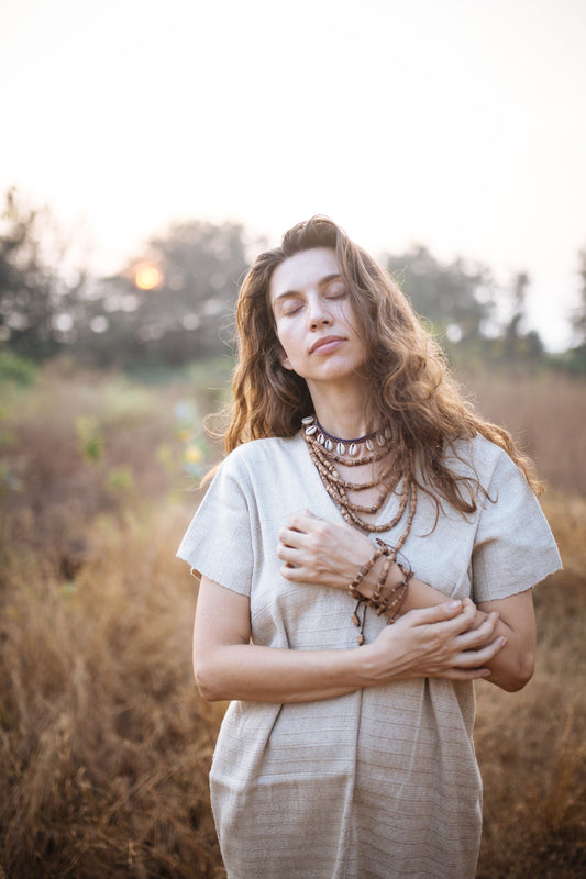 Close-up of a woman in the Karen Hill Tribe hemp kaftan with eyes closed, hands resting over her heart. The natural
cream-colored handwoven fabric texture is visible up close, paired with wooden bead bracelets. Outdoor setting with
golden grass in the background.