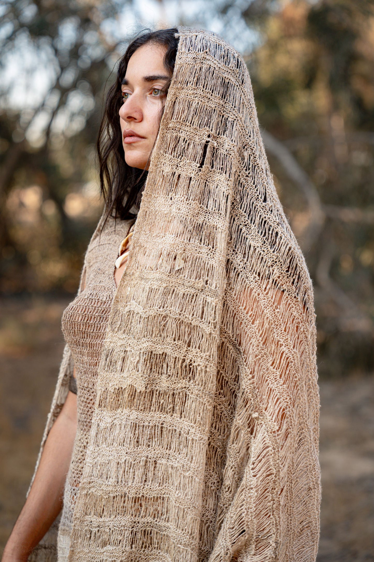 Close-up of a model wrapped in a thin natural nettle shawl with an airy open weave and striped texture, photographed in nature