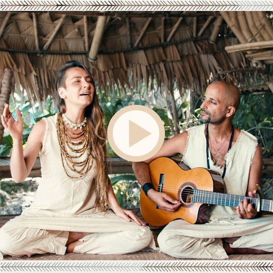 Olga singing while Yinon plays acoustic guitar, seated cross-legged in a bamboo hut wearing boho hippie clothing in natural tones and raw silk.