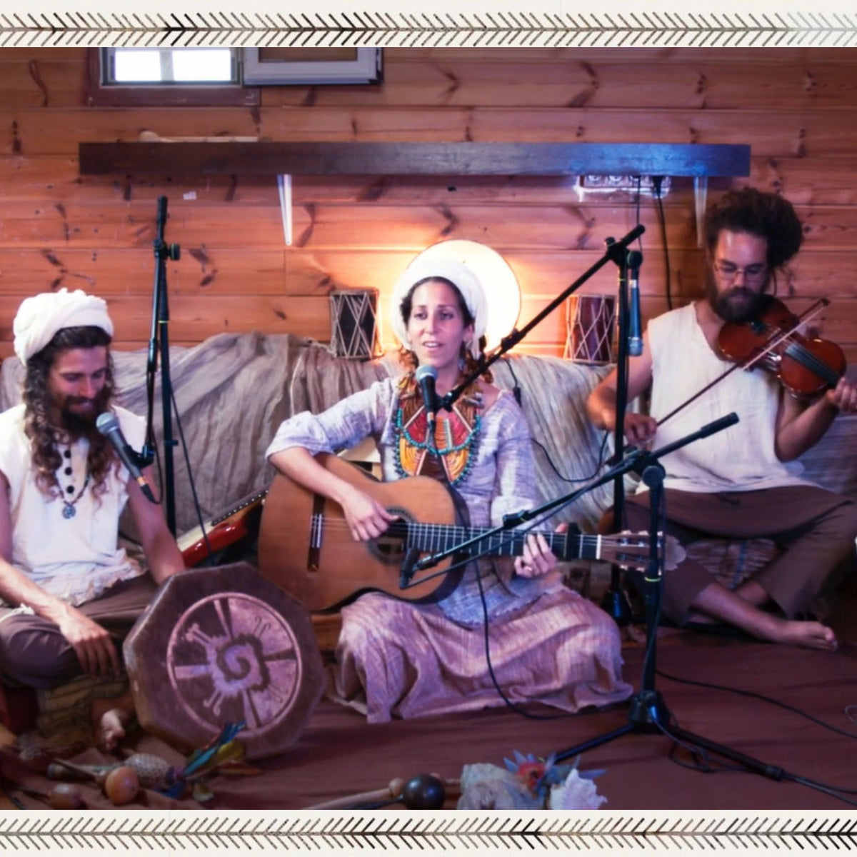 few musicians performing indoors on a wooden stage, woman singing and playing guitar, with a violinist and a drummer, wearing earthy boho clothing.