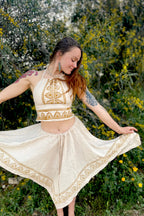 Woman wearing natural fabric crop top and flowing skirt with tribal patterns in beige and brown tones from Ancient Butterfly Outfit Skirt Top Camel Blockprint collection, standing outdoors with greenery and yellow flowers in background.