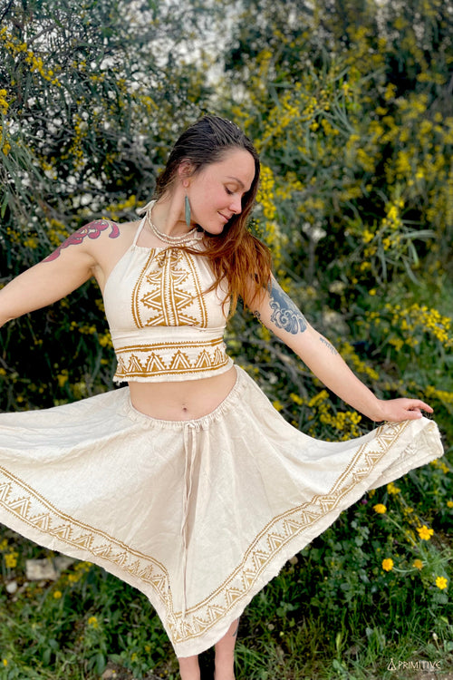 Woman wearing natural fabric crop top and flowing skirt with tribal patterns in beige and brown tones from Ancient Butterfly Outfit Skirt Top Camel Blockprint collection, standing outdoors with greenery and yellow flowers in background.