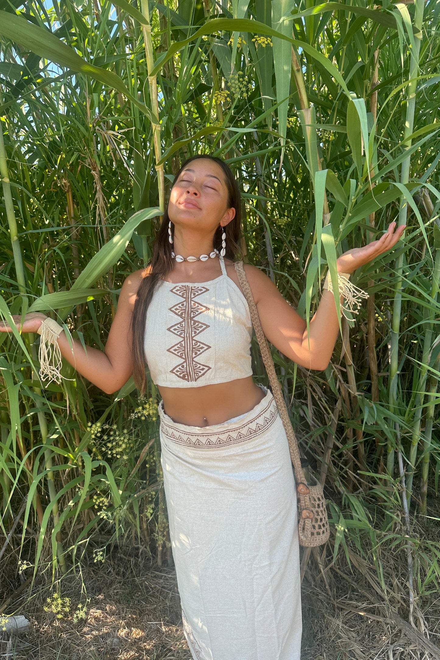 Full-body view of a Boho Woman in a raw silk halter top with tribal block print, standing among tall green reeds in natural sunlight.