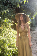 A woman wearing a wide-brimmed straw hat and a sleeveless olive-brown maxi dress with a tie waist, standing outdoors on a sunlit path with lush green foliage on one side.