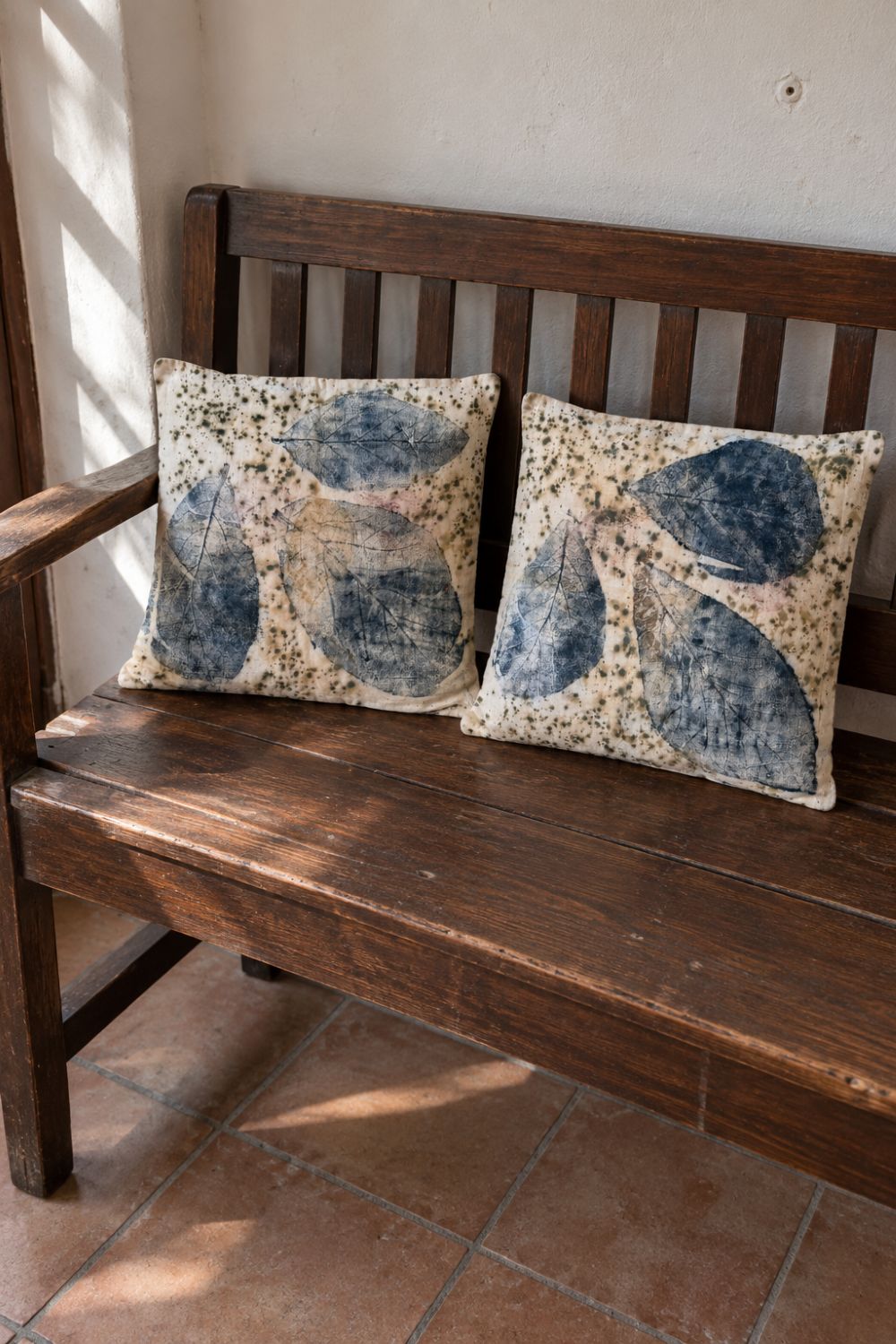 Two botanical print pillow covers in cream handloom cotton with blue leaf prints and olive green speckles, on a dark wooden bench in afternoon sunlight