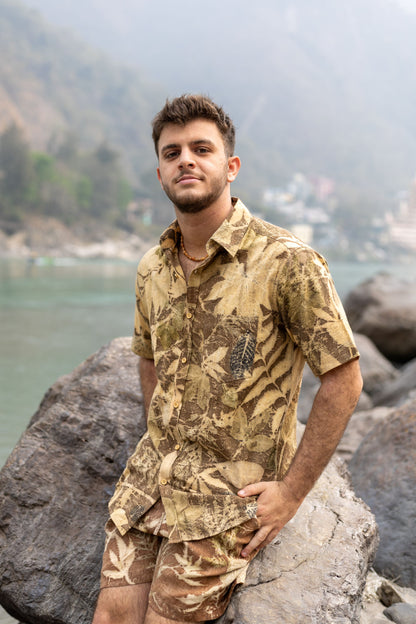 Man leaning on a rock near a river wearing an earth-toned botanical leaf print shirt and shorts.