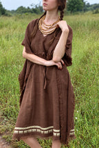 Model with braided hair wearing a brown khadi cotton kaftan in a grassy field, showing its textured fabric, fringed edges and cream block-printed hem.
