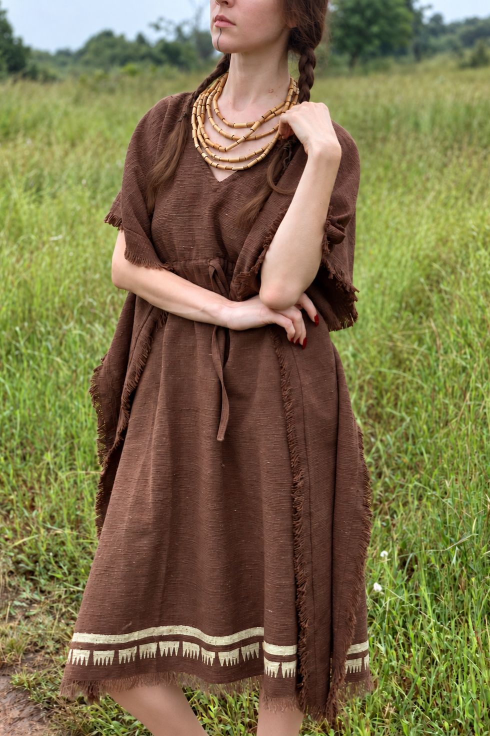 Model with braided hair wearing a brown khadi cotton kaftan in a grassy field, showing its textured fabric, fringed edges and cream block-printed hem.