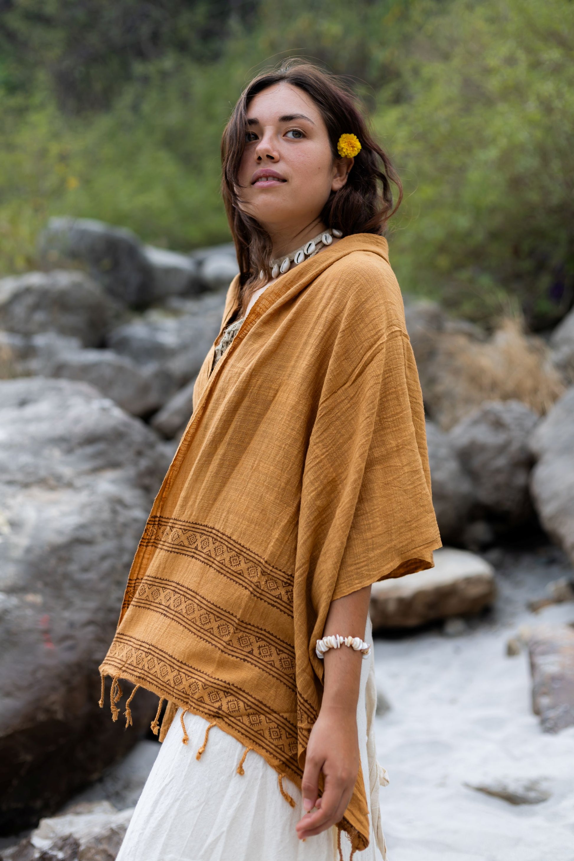 Woman wearing a Brown shawl with intricate patterns, standing in a natural setting with rocks and greenery.