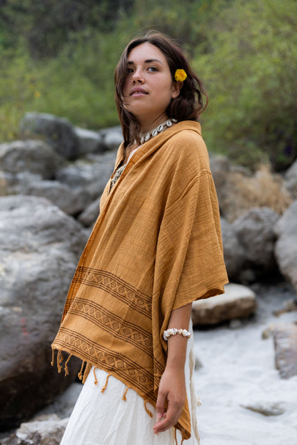 Woman wearing a Brown shawl with intricate patterns, standing in a natural setting with rocks and greenery.