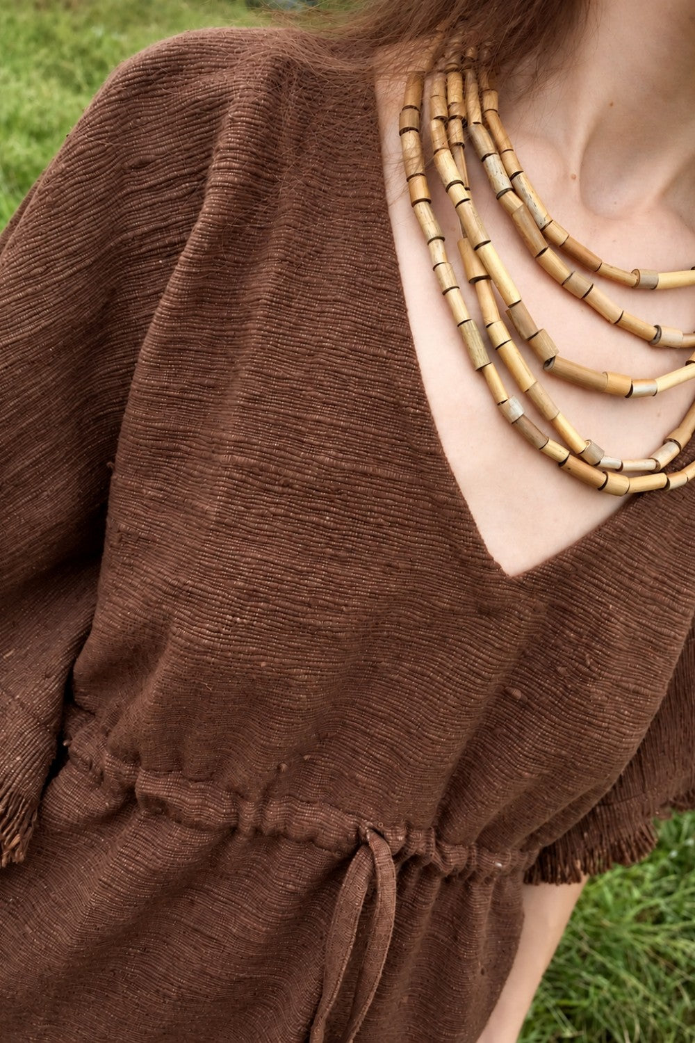 Brown khadi cotton kaftan showing textured weave, V-neckline, fringed sleeve and waist drawstring, worn with a layered bead necklace.