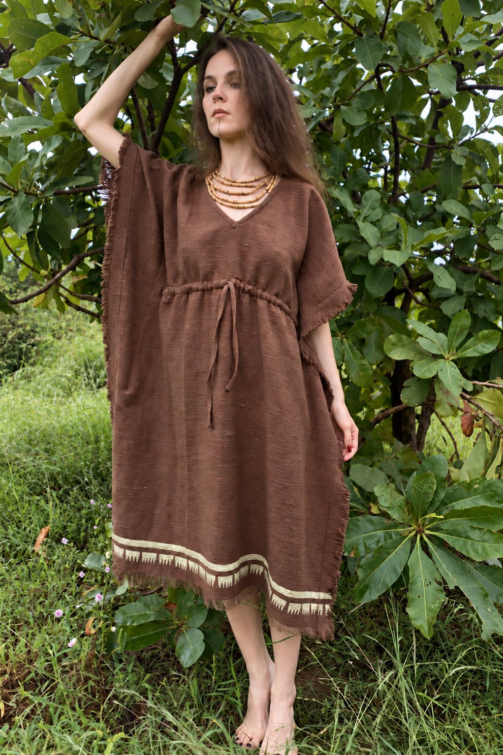 Full-length view of a model beneath a tree wearing a brown khadi cotton kaftan with wide sleeves, an adjustable waist and cream block prints along the hem.
