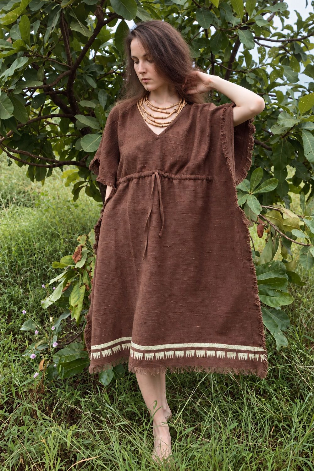 Front view of a brown khadi cotton kaftan with a drawstring waist, V-neck and cream block-printed border, worn in a leafy garden.