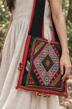 Close-up of a black shoulder bag with colorful geometric embroidery, red trim, a wooden button, and small multicolored tassels, worn over a white linen dress.