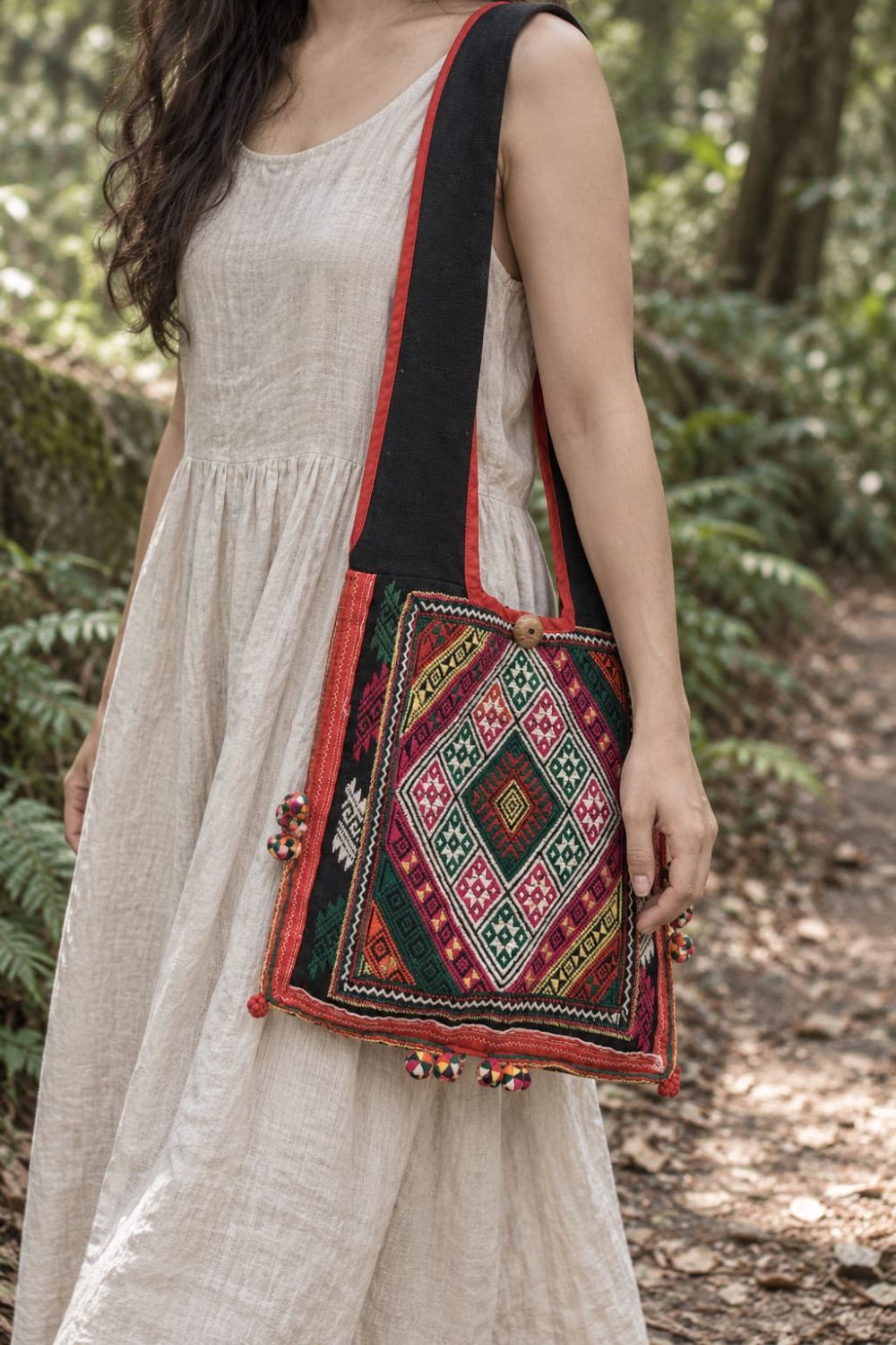 Woman in a white linen dress carrying a colorful embroidered black shoulder bag with a wide strap and red trim on a leafy woodland path.
