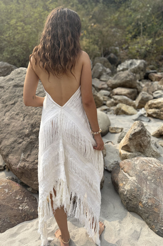 Woman in a white fringe dress standing on rocky terrain with greenery in the background