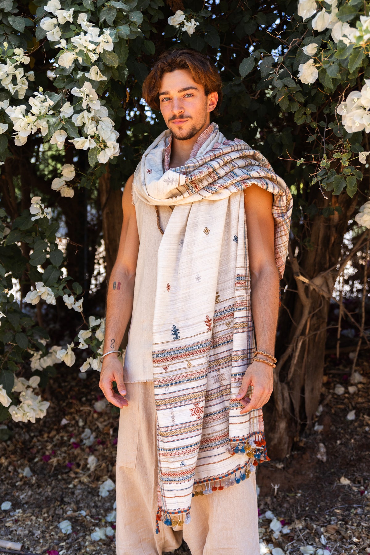 Man wearing an oversized Gujarati tribal shawl with colorful geometric motifs and tassel edge, styled as a draped wrap outdoors among white flowers.