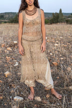 Front view of a sleeveless hand-knitted nettle dress in natural undyed greige, with a leaf-and-chevron lace pattern and a drop-waist band. It’s worn over loose beige harem pants with wooden bead necklaces, barefoot in a dry field.