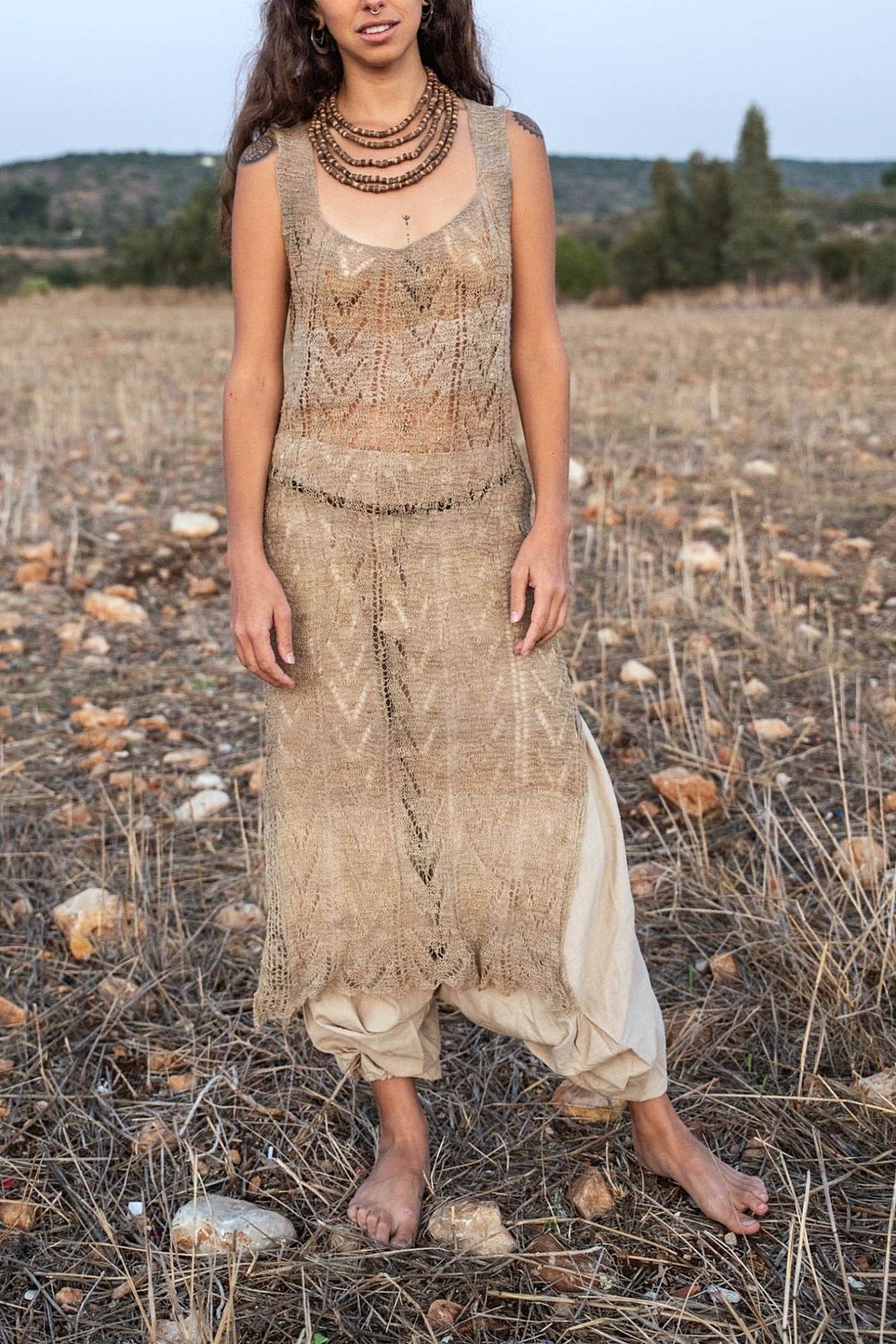 Front view of a sleeveless hand-knitted nettle dress in natural undyed greige, with a leaf-and-chevron lace pattern and a drop-waist band. It’s worn over loose beige harem pants with wooden bead necklaces, barefoot in a dry field.