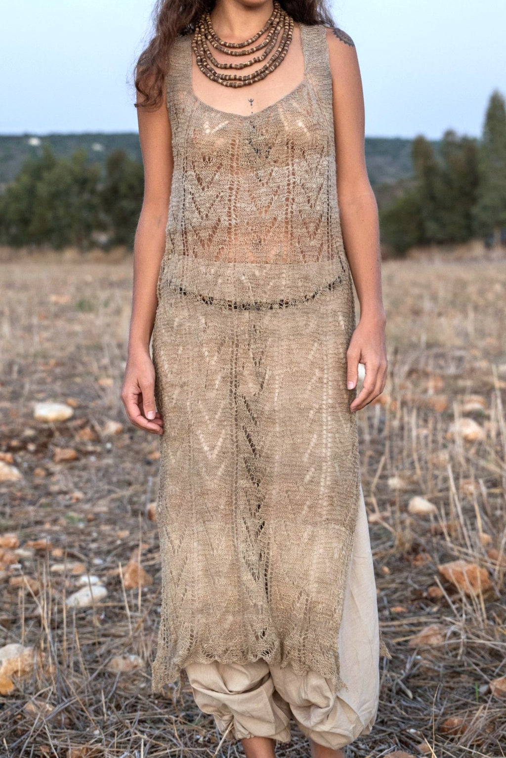 Close front view of a long hand-knitted nettle lace dress showing its open chevron stitch pattern, scoop neck and drop waist. It’s layered over beige harem pants and styled with stacked wooden bead necklaces.