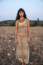 Model standing in a dry field at dusk in a sleeveless, ankle-length hand-knitted nettle dress with a sheer lace pattern, worn over beige harem pants, with layered wooden bead necklaces.