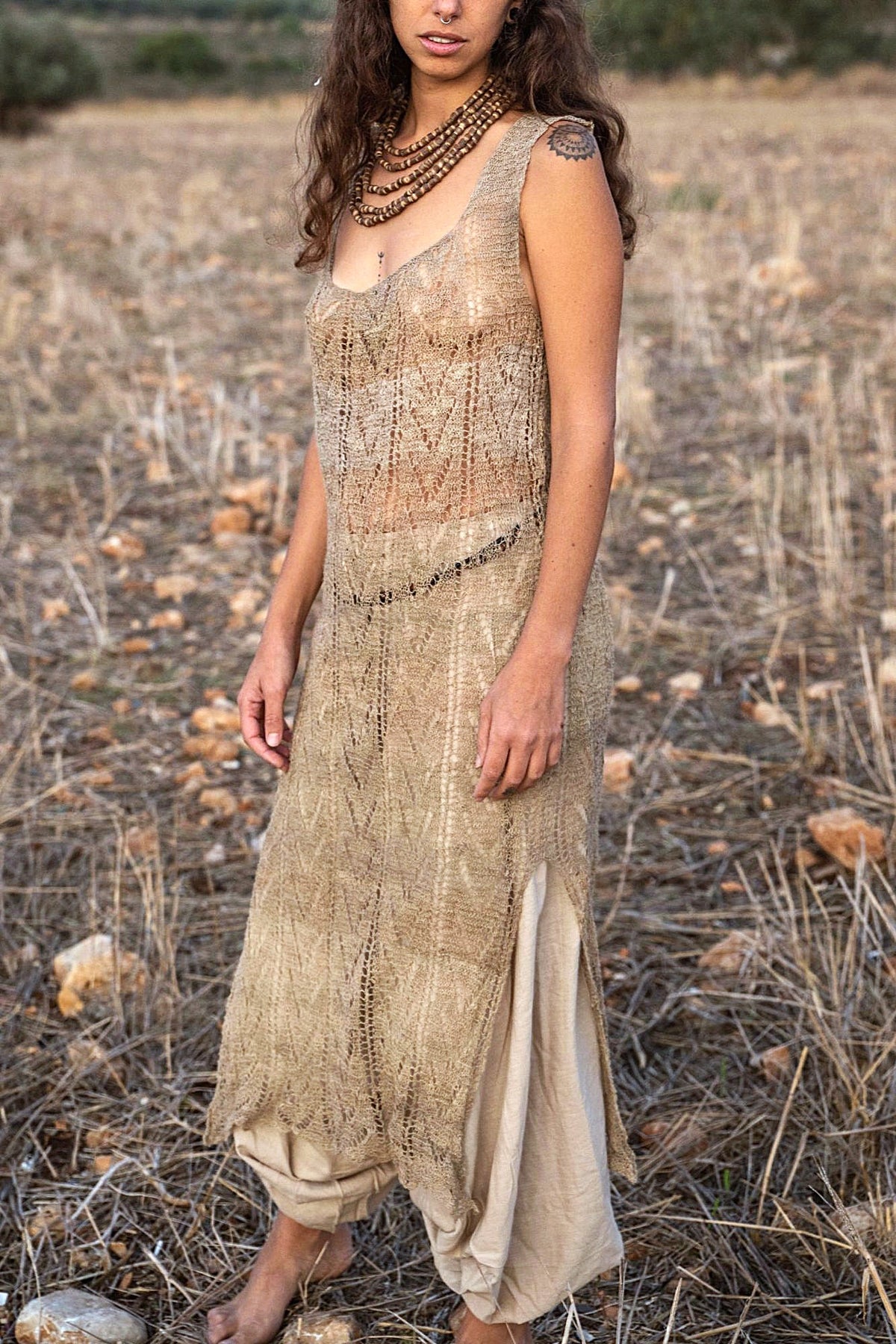 hand-knitted nettle lace dress in natural greige, showing the side slit with beige harem pants underneath, the scalloped hem and the open-knit chevron pattern.