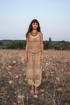 Full-length front view of a model smiling in a natural undyed hand-knitted nettle lace dress worn over gathered beige harem pants, barefoot in a dry field at dusk.
