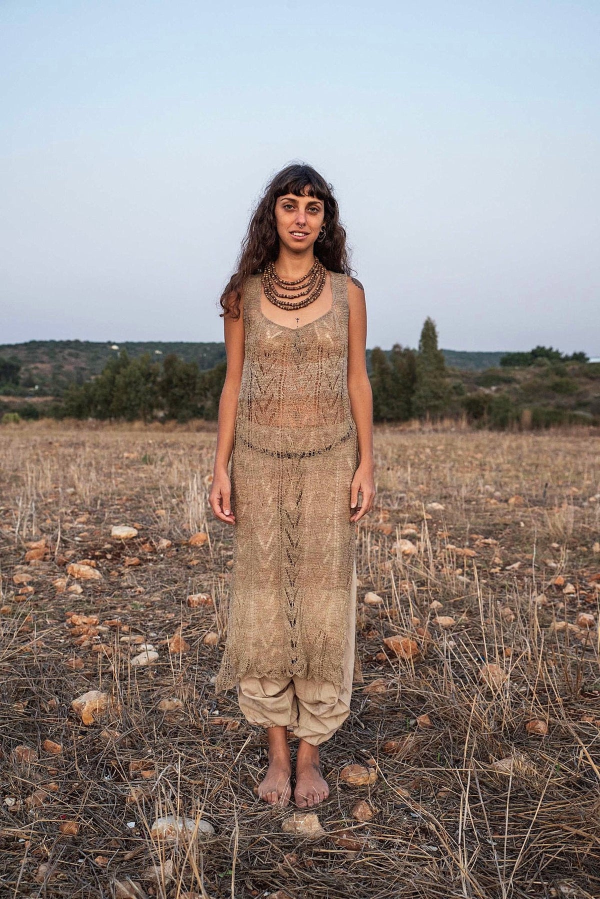Full-length front view of a model smiling in a natural undyed hand-knitted nettle lace dress worn over gathered beige harem pants, barefoot in a dry field at dusk.