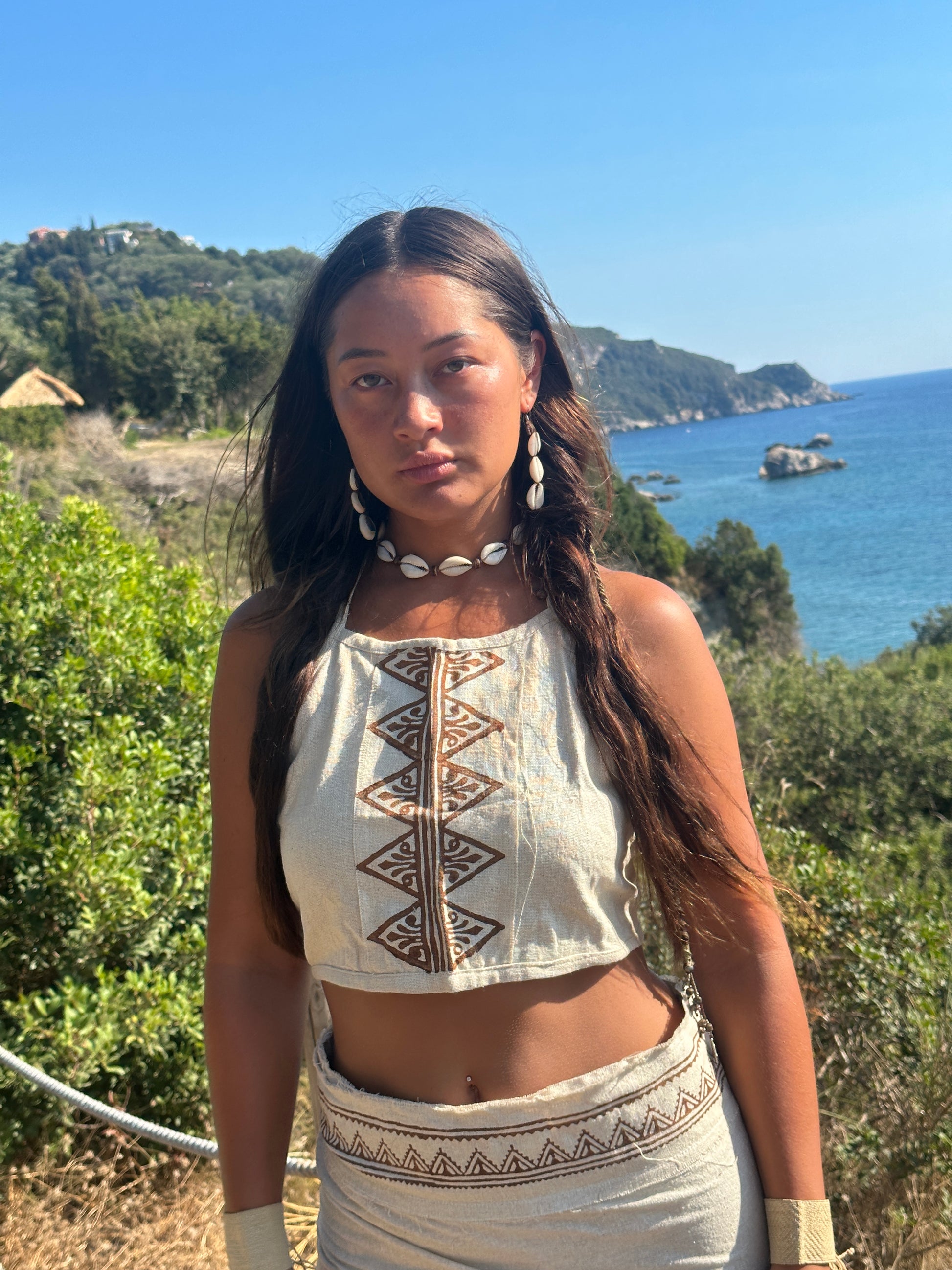 Hippie woman wearing a natural raw silk halter crop top with tribal block print, photographed by the sea with coastal cliffs in the background.