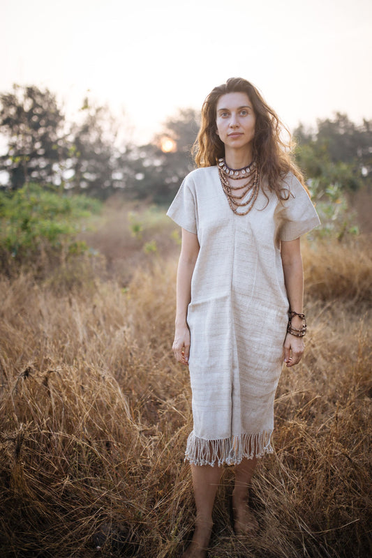 wearing a Karen Hill Tribe handwoven hemp kaftan in natural color, standing in a golden grass field at sunset.
The knee-length dress features short sleeves, a V-neckline, and decorative fringe along the hem.
