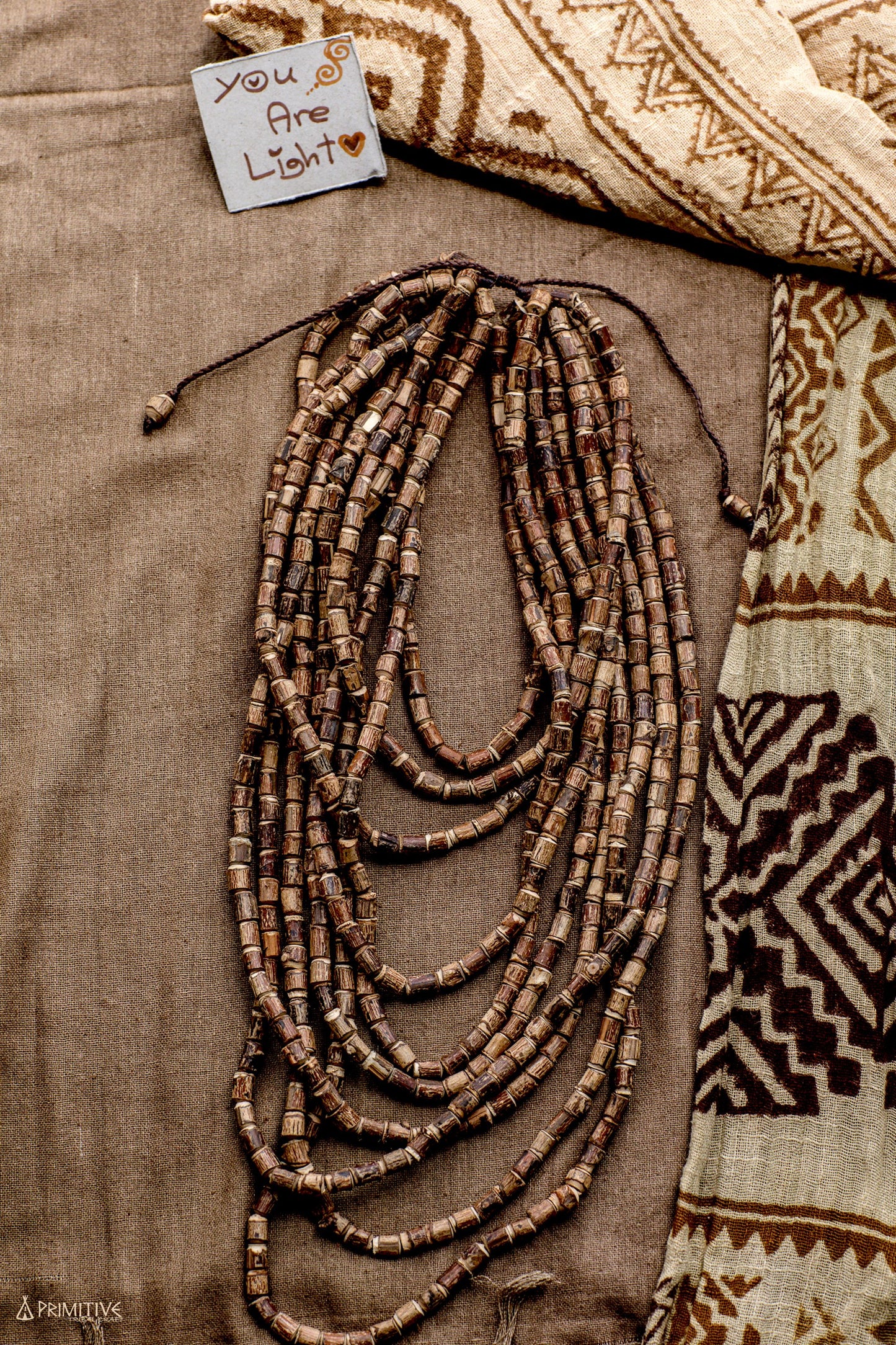 Flat lay of the 10-strand Tulsi wood beads necklace arranged on a patterned tribal textile, showing the full spread of hand-cut unpolished beads and natural brown wood tones