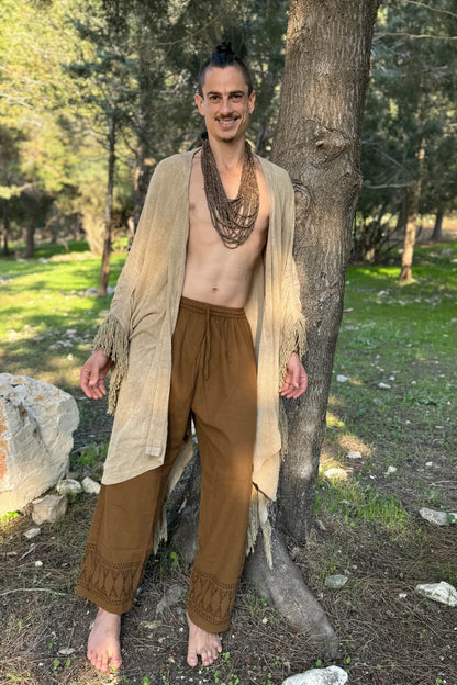 A man standing in a forest wearing brown loose straight-leg cotton pants featuring a tribal tipi block print at the ankles