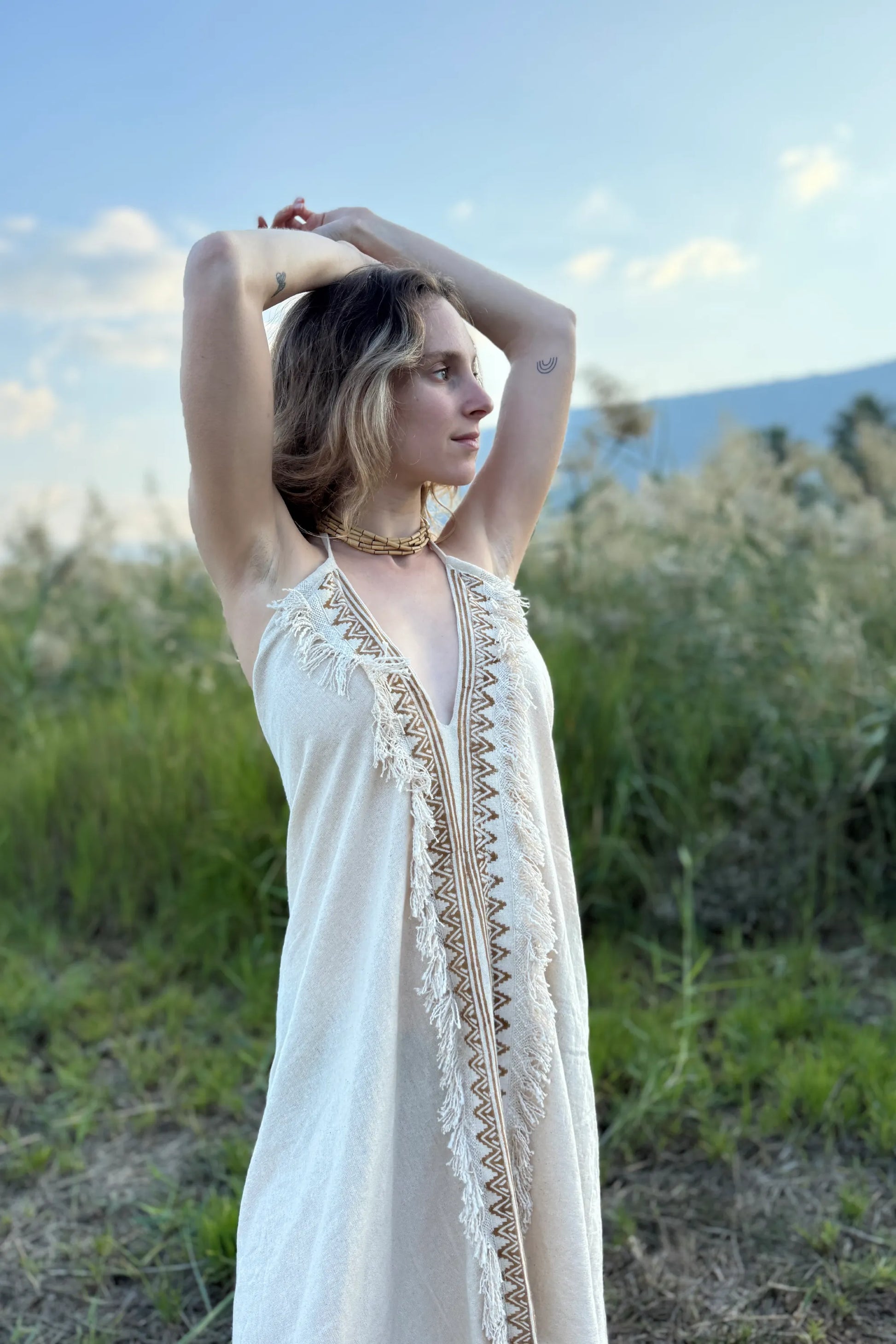 Woman in a short cotton butterfly dress with sleeveless cut, fringed edges and patterned front, standing in a field with a blue sky.