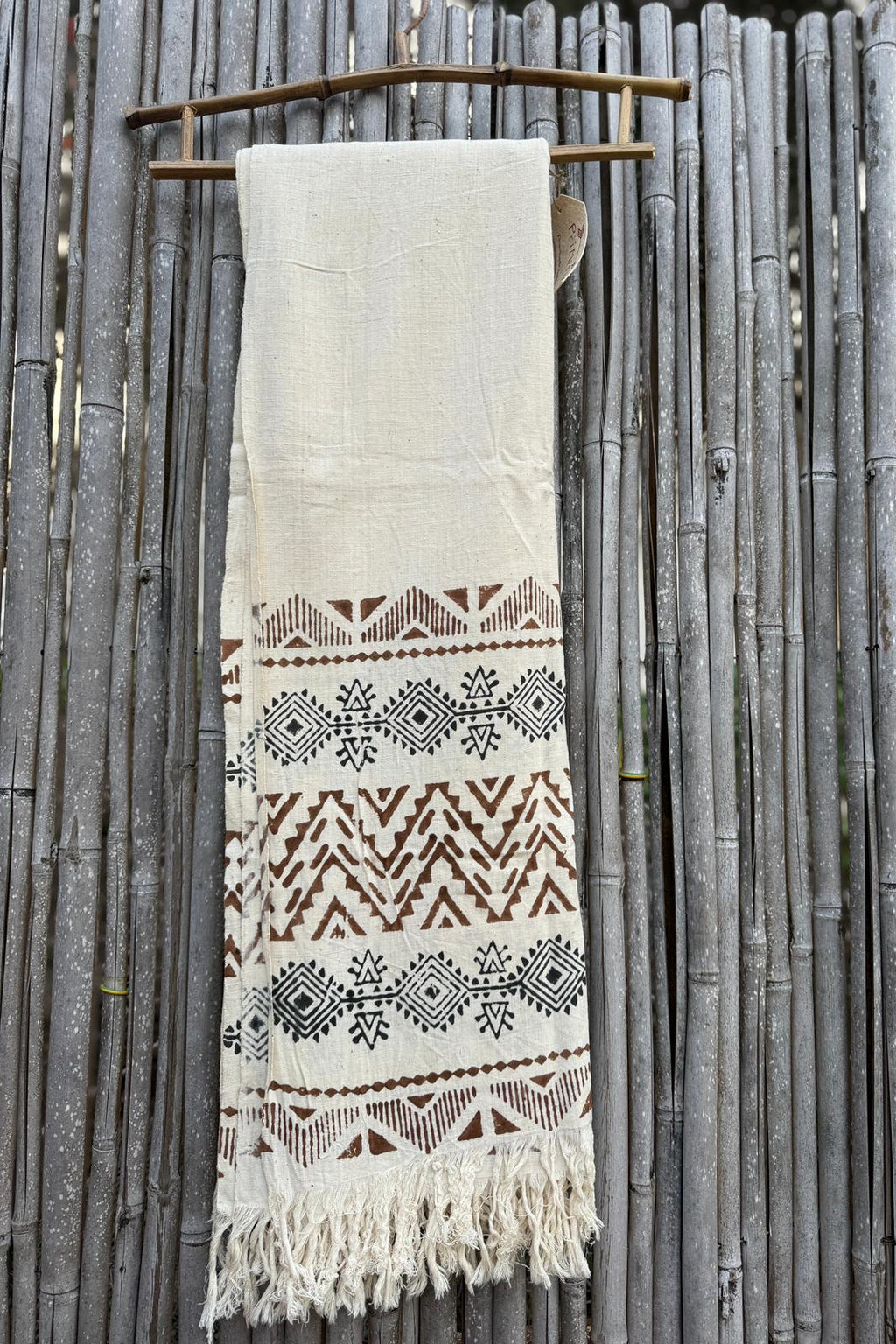 Natural khadi cotton scarf with brown tribal pattern bands and fringe, photographed hanging on a bamboo fence.