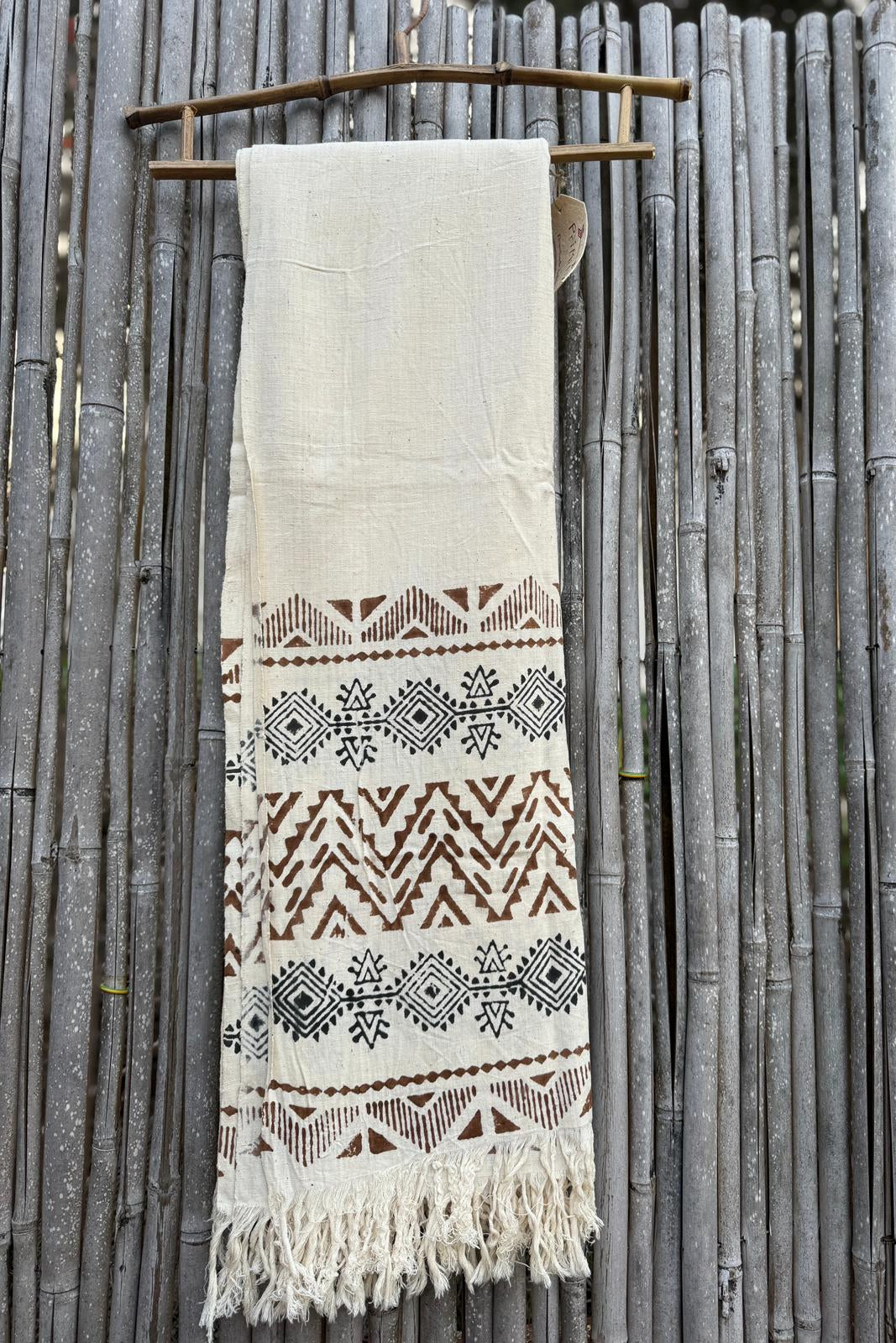 Natural khadi cotton scarf with brown tribal pattern bands and fringe, photographed hanging on a bamboo fence.