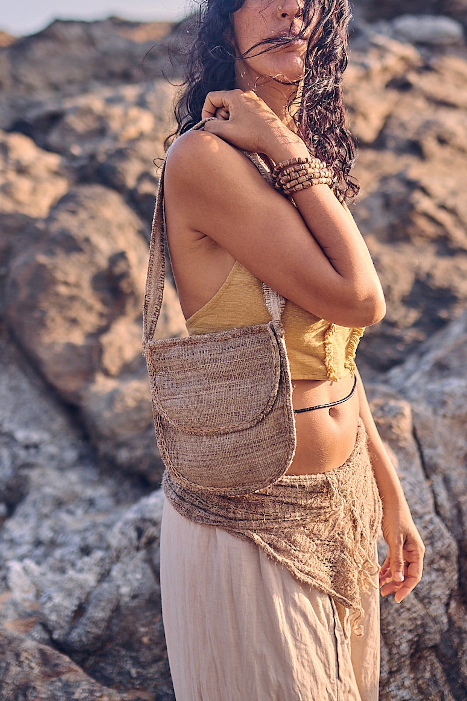 Woman carrying a handstitched wild nettle and hemp side bag over her shoulder, standing by sunlit coastal rocks, wearing natural earthy clothing