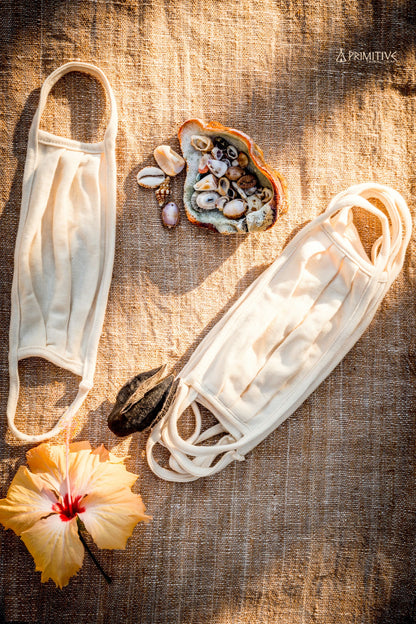 Two white Organic Cotton face masks on a textured surface with a shell, flowers, and pebbles.