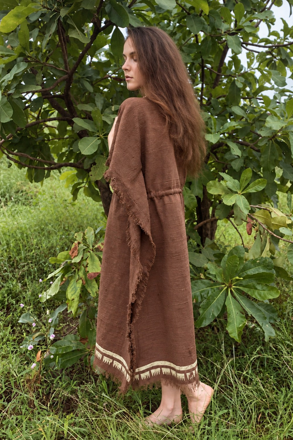 Model wearing a brown khadi cotton kaftan with loose sleeves, fringed edges and cream block-printed hem, viewed from the side among green trees.
