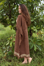 Model wearing a brown khadi cotton kaftan with loose sleeves, fringed edges and cream block-printed hem, viewed from the side among green trees.