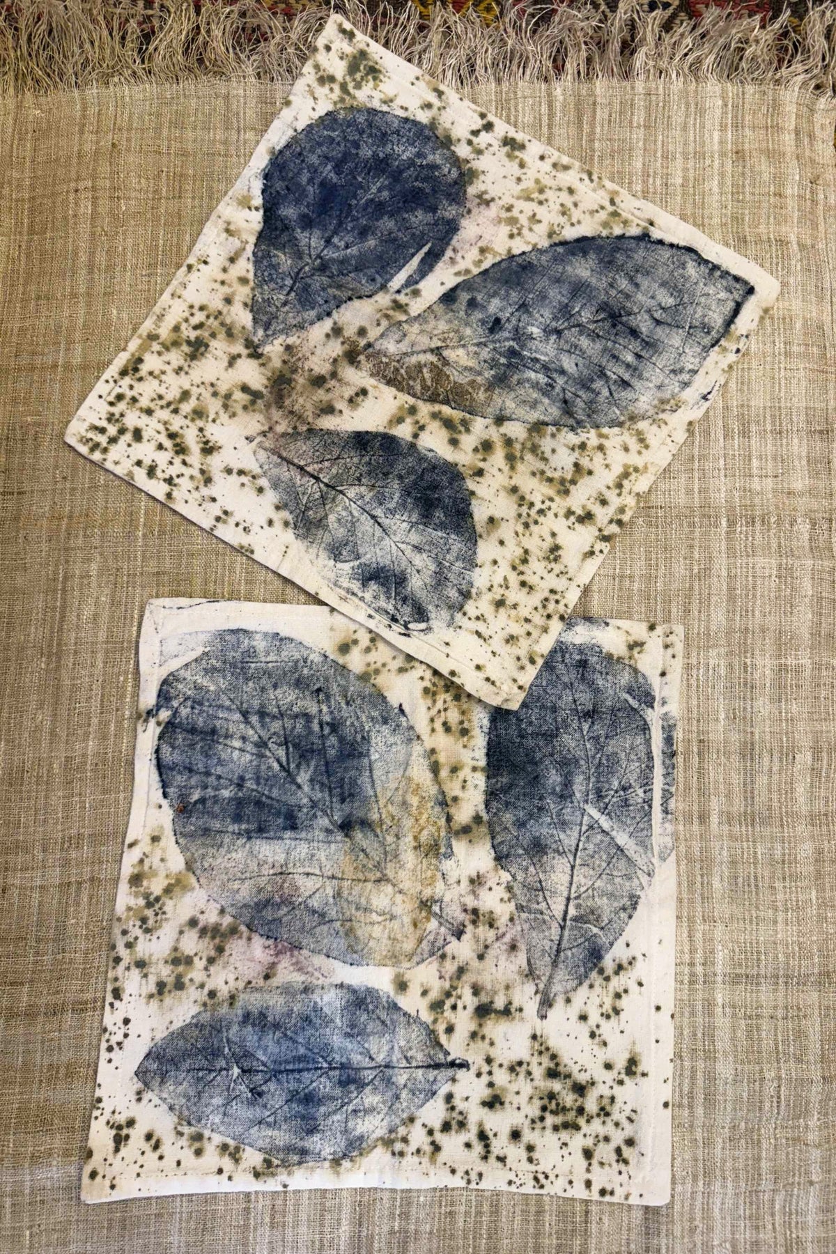 Two flat botanical print pillowcases with blue leaf vein prints, soft pink tones and olive speckles on cream cotton, laid on natural handwoven fabric
