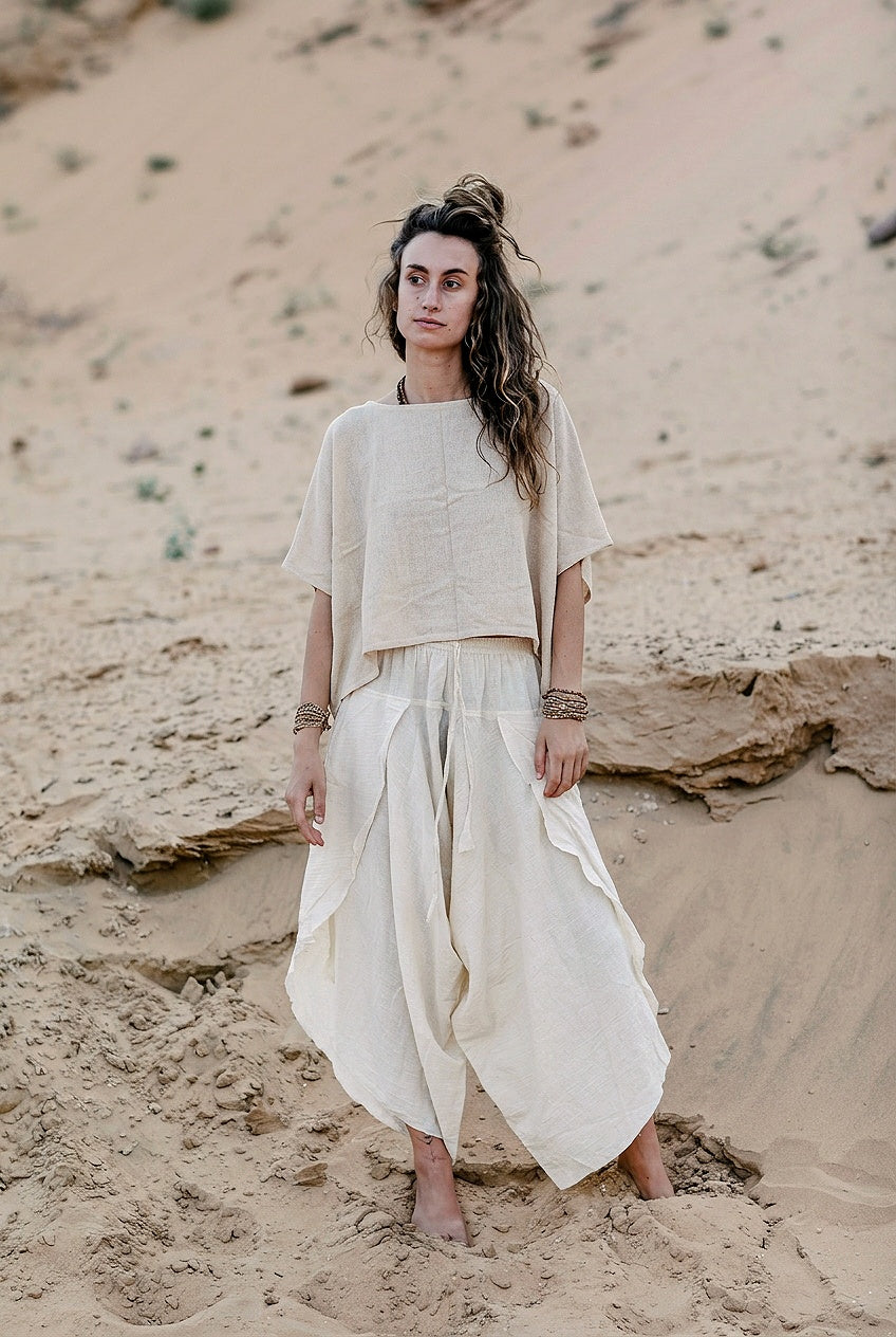 A woman standing barefoot in sand, modeling a nomadic-style two-piece outfit. The set features a loose, wide-neck raw silk top and matching flowy, asymmetrical pants in a natural, unbleached beige tone.