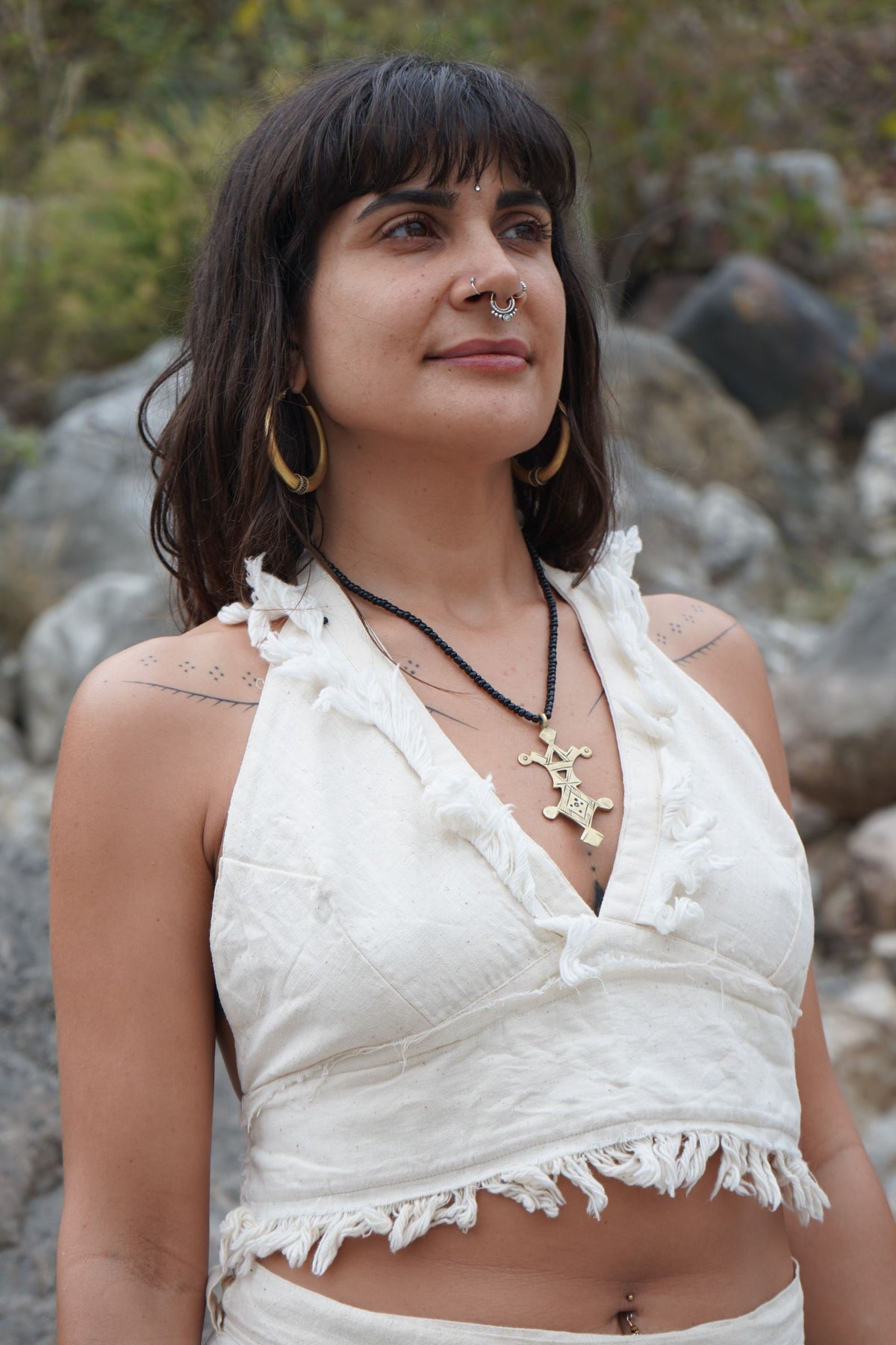 Woman wearing a white top with fringe details, standing outdoors with rocks and greenery in the background.