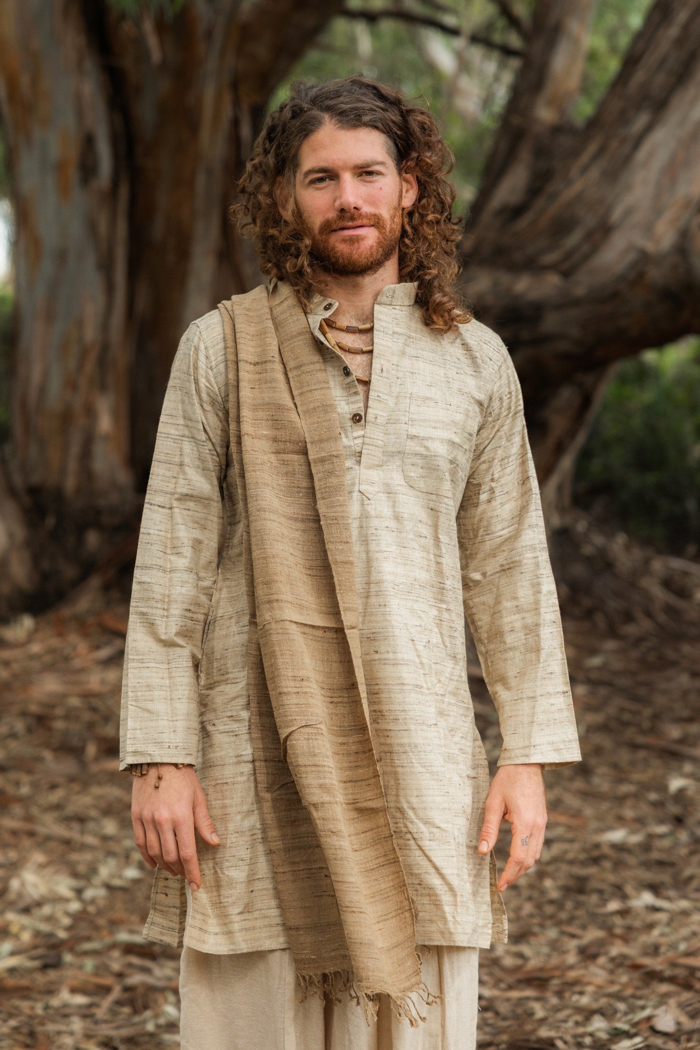 Man wearing a natural wild silk traditional long kurta open at the collar with a beaded necklace, outdoors among trees