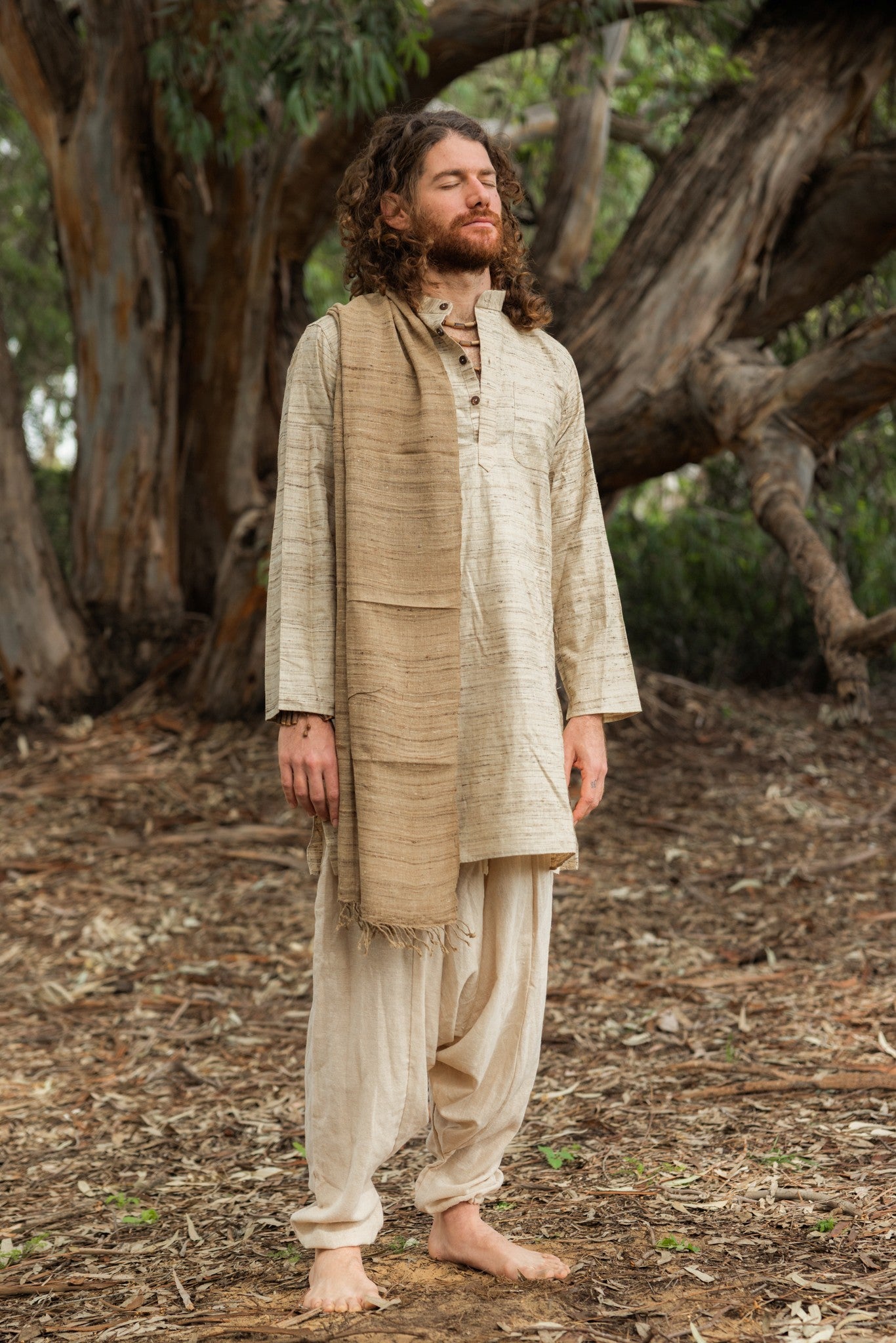 Man with eyes closed wearing a full-length natural wild silk kurta, standing peacefully in front of a large tree