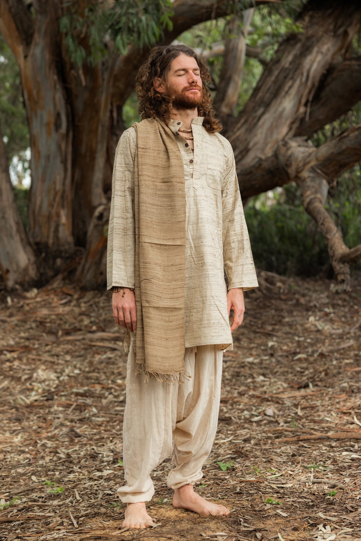 Man with eyes closed wearing a full-length natural wild silk kurta, standing peacefully in front of a large tree