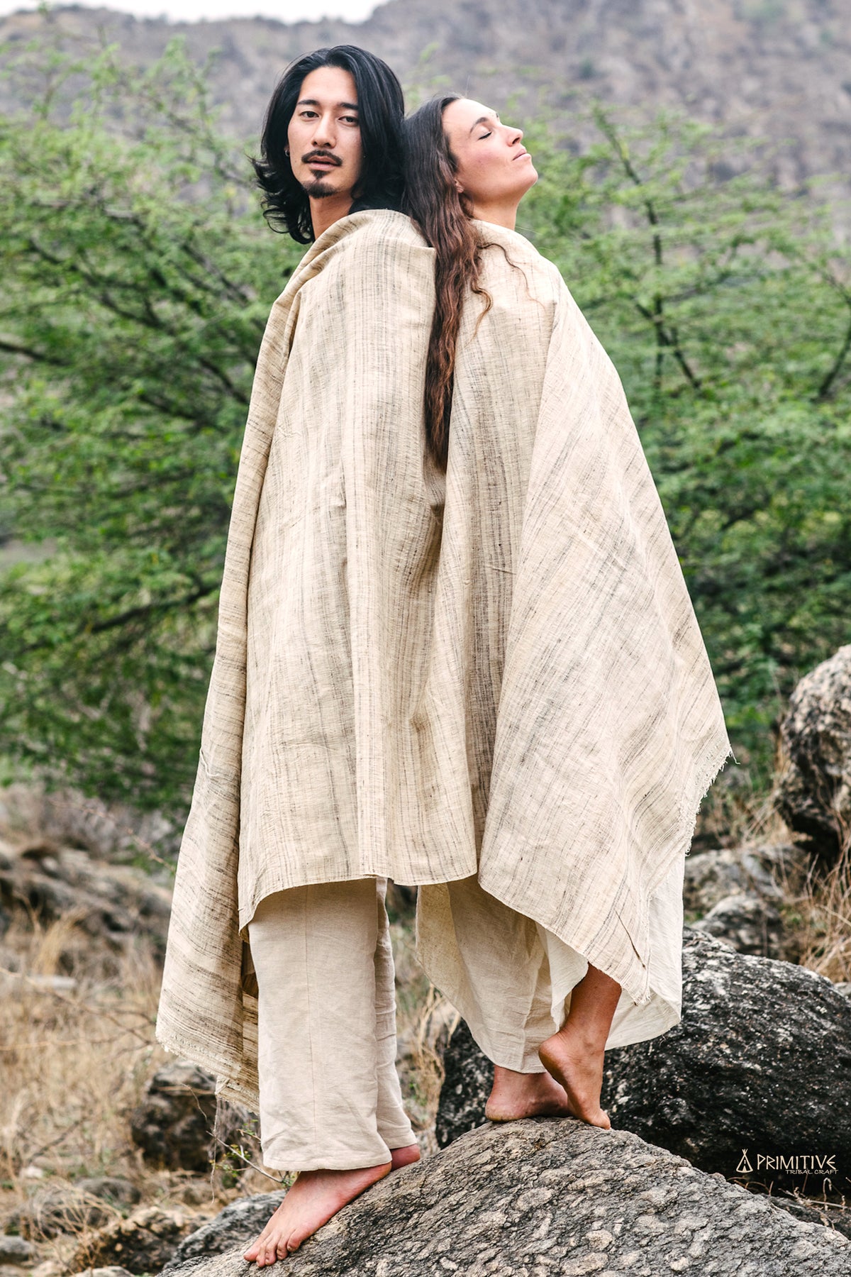 Two individuals standing on rocks, draped in beige handwoven tribal shawls, with a natural landscape in the background.