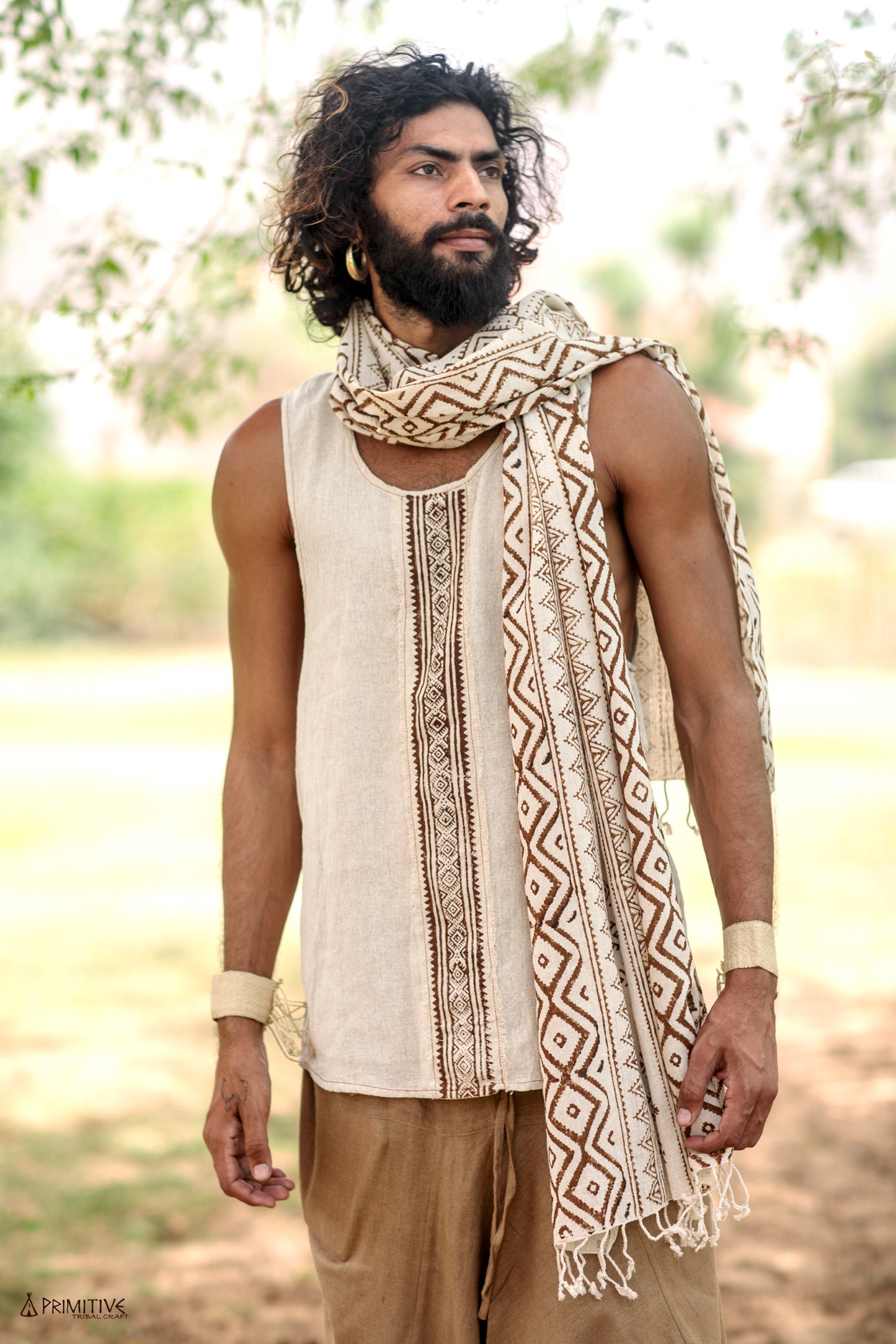 A boho man wearing a traditional beige handwoven raw silk tank top with brown block print patterns, paired with a white scarf, standing outdoors.