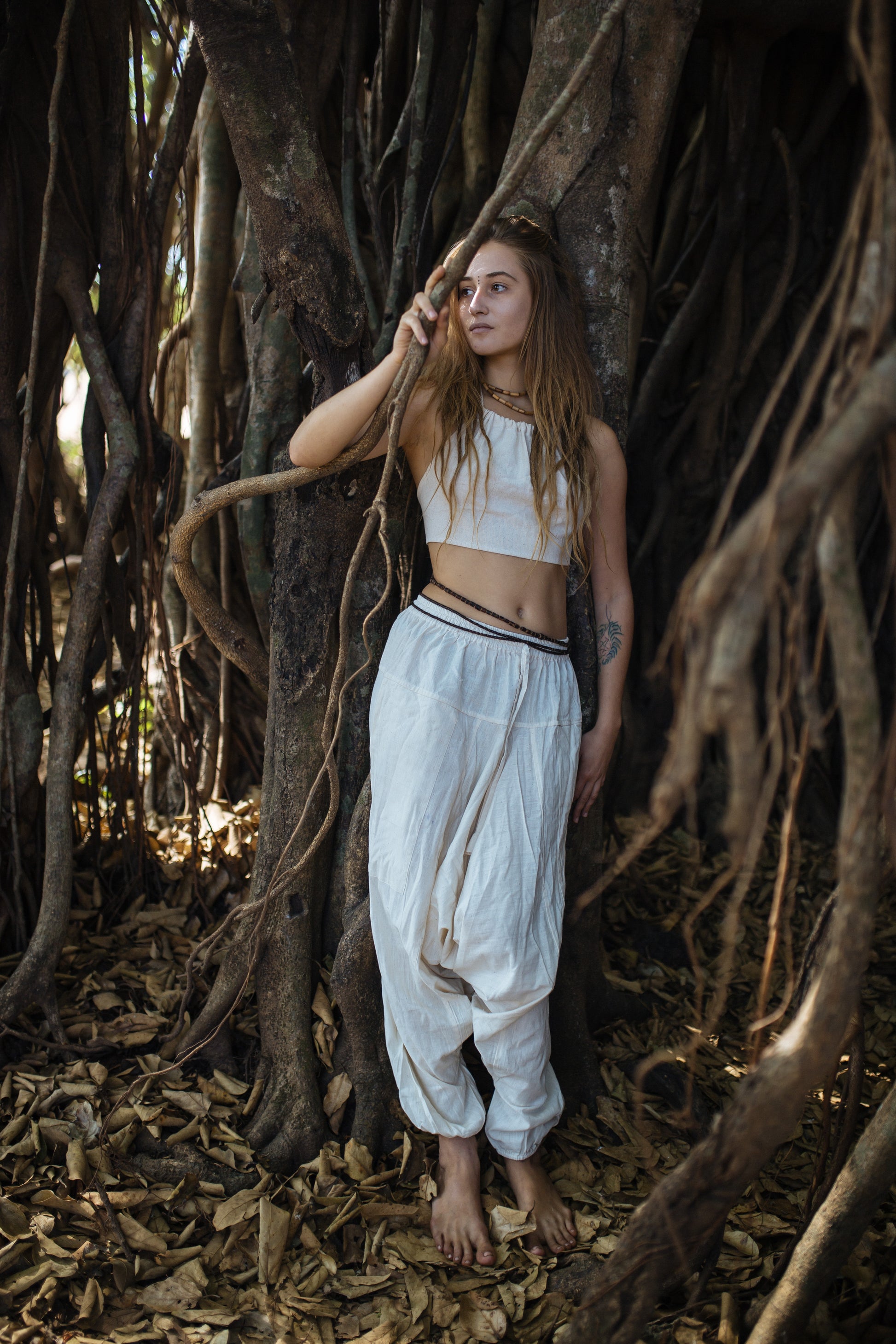 Woman wearing neutral natural cotton harem pants with drop-crotch relaxed fit and drawstring waist, styled for festival dance and yoga in a forest setting.