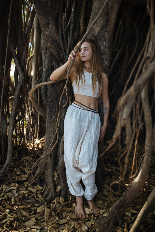 Woman wearing neutral natural cotton harem pants with drop-crotch relaxed fit and drawstring waist, styled for festival dance and yoga in a forest setting.
