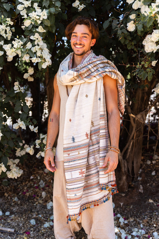 Smiling model styled in a large handwoven Gujarati embroidered shawl with patterned borders and tassels, wrapped over neutral outfit in a leafy outdoor setting.